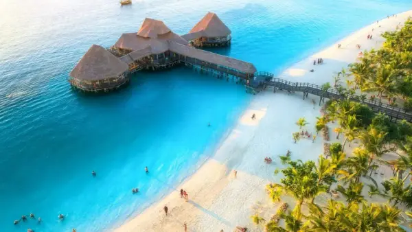 Zanzibar w czerwcu: idealna pogoda na wypoczynek na rajskiej plaży z turkusową wodą i palmami. Widać hotel na wodzie i ludzi kąpiących się.