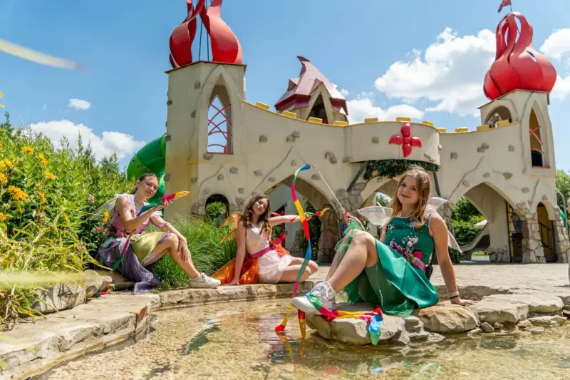 Trzy wróżki w kolorowych strojach siedzą przy strumyku przed bajkowym zamkiem. To z pewnością jedne z wielu magiczne ogrody atrakcje.